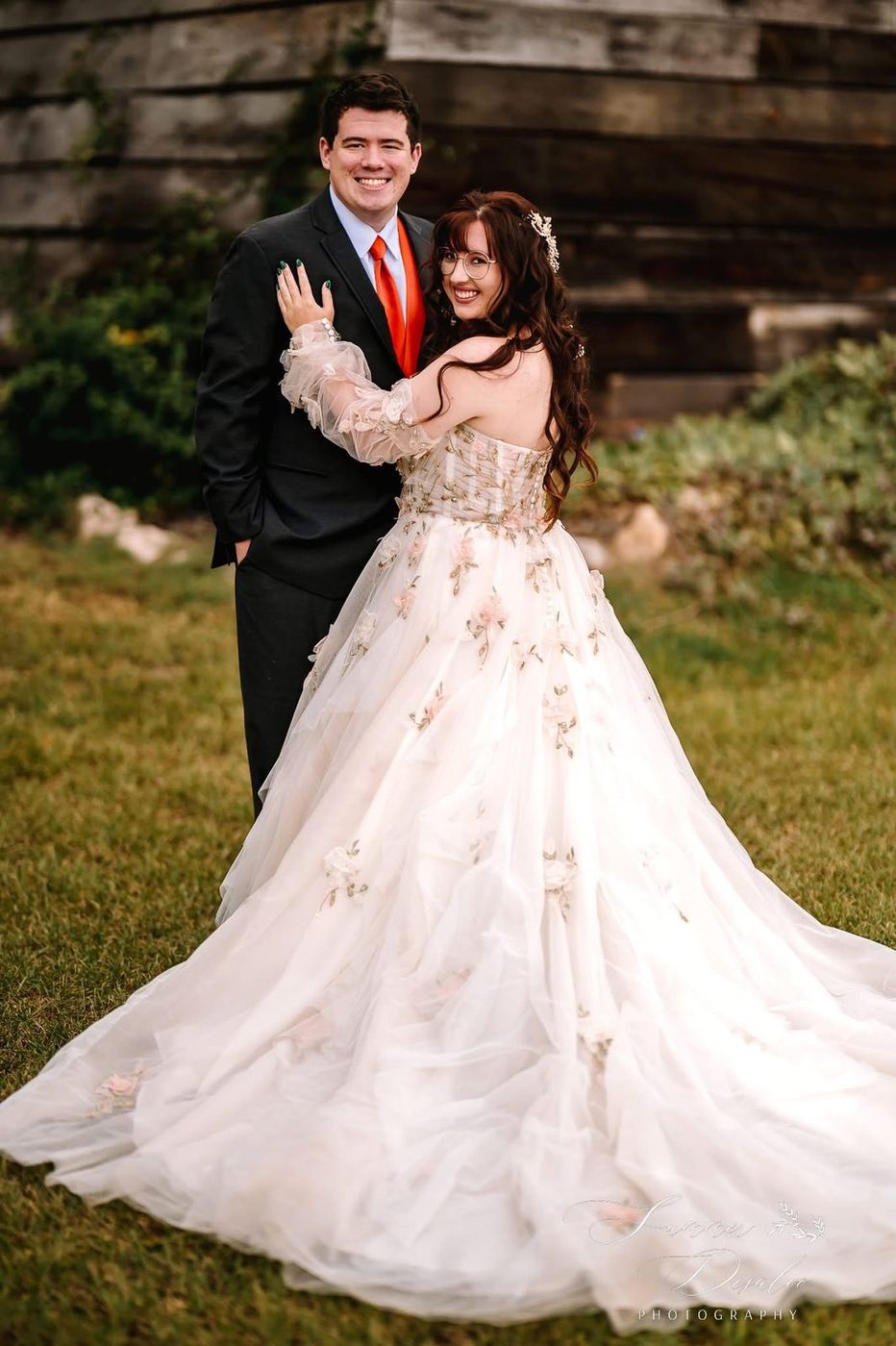 Bride in a floral tulle ball gown with the groom in a charcoal suit and orange tie (photographer watermark shown)