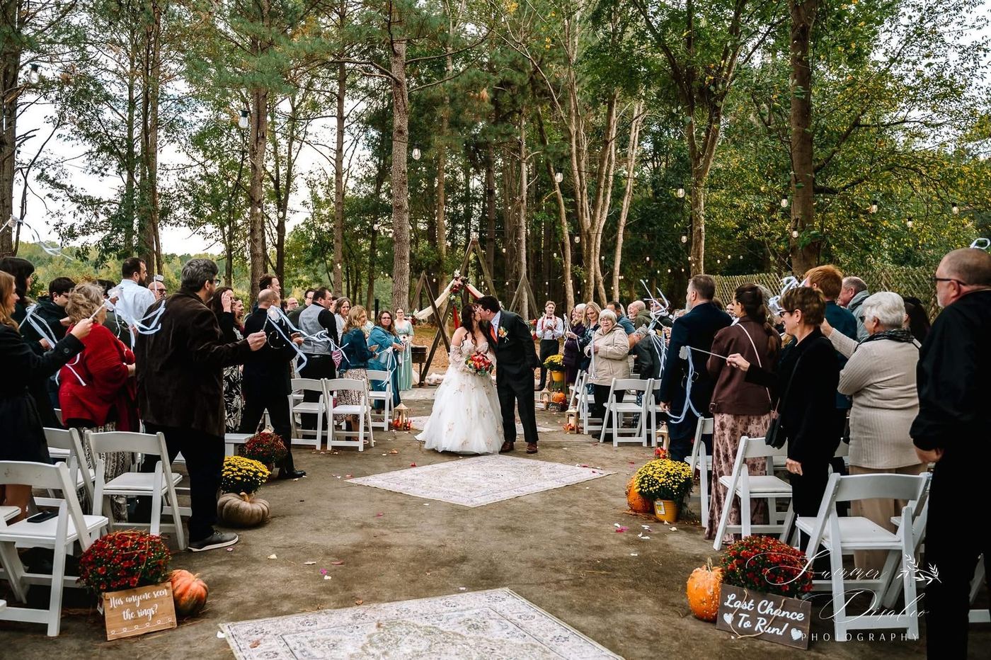 First kiss at a woodland ceremony with ribbon wands, mums, pumpkins and rugs lining the aisle (photographer watermark shown)